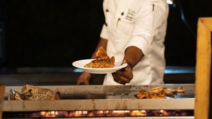 A chef dressed in white presenting a plate of grilled food with the sea in the background - Kaldan Samudra Palace